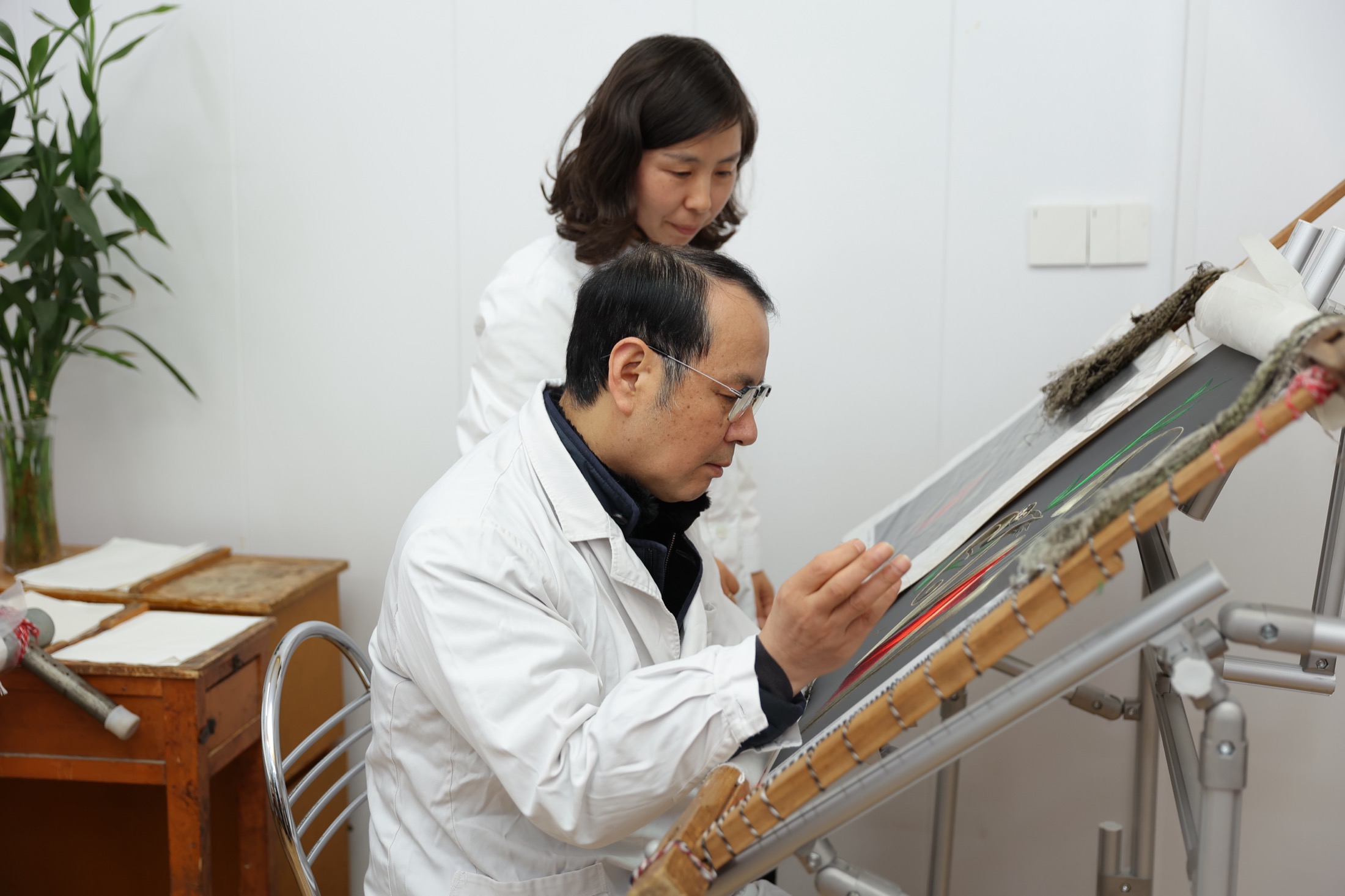 两位刺绣工作室人员在倾斜织架前查看丝绣作品。